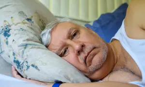 Starszy mężczyzna leży w łóżku, zamyślony. Jego twarz wyraża niepokój, być może związane z cichym udarem i jego objawami.