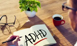 Osoba zmagająca się ze spektrum ADHD pisze w zeszycie. Obok leżą okulary, filiżanka kawy i roślina.