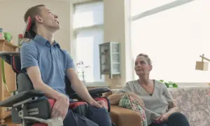 Młody mężczyzna w wózku inwalidzkim, z widocznymi **sla objawy**, uśmiecha się, patrząc w górę. Obok siedzi kobieta.