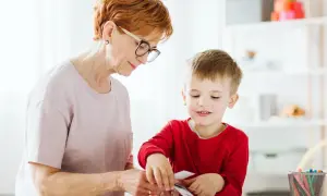 Babcia i wnuczek tworzą coś razem. Dziecko z zaciekawieniem przygląda się swoim ruchom, a jego manieryzmy ruchowe są widoczne w skupieniu.
