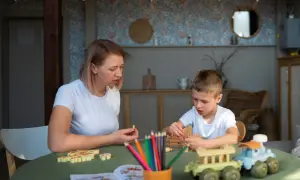 Mama i syn bawią się drewnianymi klockami i kredkami. Tematyka zabawy nawiązuje do przedszkola, rozwijając kreatywność i wyobraźnię.