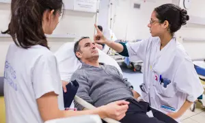 Lekarka bada pacjenta leżącego w łóżku szpitalnym, drugą pielęgniarkę widać z boku.