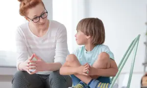 Kobieta z okularami i chłopiec siedzą naprzeciwko siebie, rozmawiając. To przykład, jak rozmawiać z dzieckiem autystycznym, budując zaufanie.