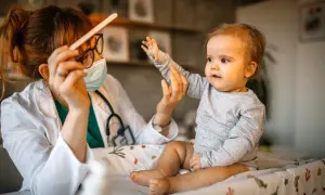 Lekarka w masce i białym fartuchu trzyma patyczek, a dziecko robi dziwne miny, wyciągając rączkę.