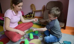 Terapia behawioralna autyzm: chłopiec i terapeutka bawią się kubeczkami na kolorowym dywanie.