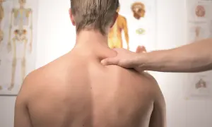 Fizjoterapeuta bada plecy pacjenta, szukając oznak zapalenia rdzenia kręgowego. Na ścianie wiszą anatomiczne plakaty.