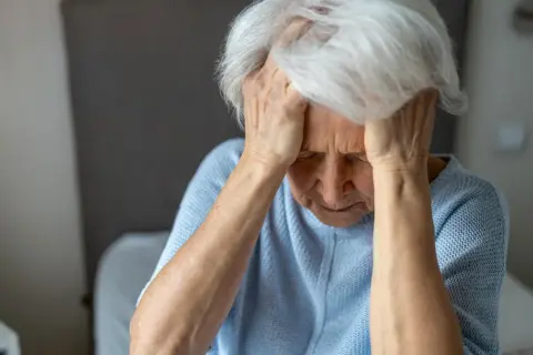 Starsza kobieta z rękami na skroniach, zamyślona, być może doświadcza pierwszych objawów demencji.
