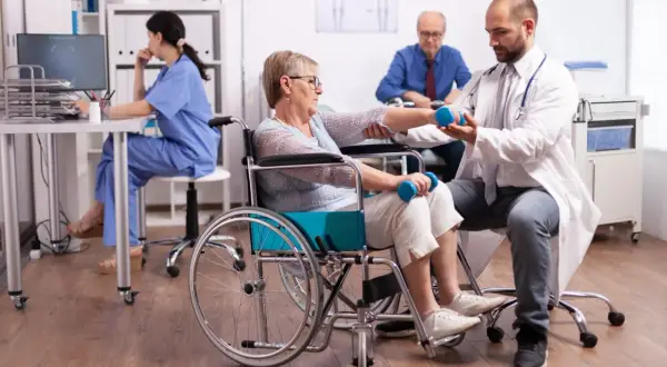 Rehabilitacja po udarze krwotocznym. Lekarz pomaga pacjentce w wózku inwalidzkim ćwiczyć z hantlami, poprawiając rokowania.