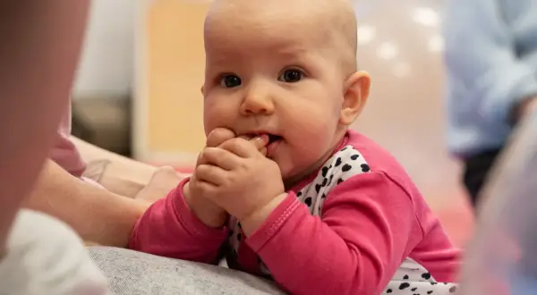 Malutkie dziecko w różowej bluzeczce z serduszkami, z paluszkami w buzi, delikatnie buja się w przód i tył, odkrywając świat.