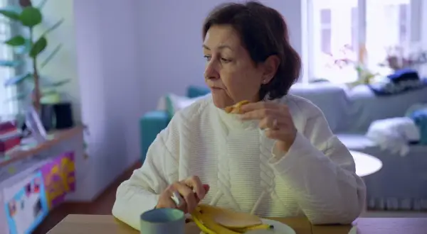 Kobieta w białym swetrze je banana, czując uczucie guli w gardle. Na stole kubek i talerz.