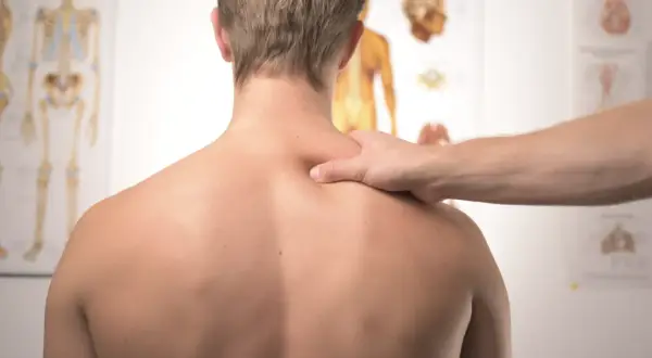 Fizjoterapeuta bada plecy pacjenta, szukając oznak zapalenia rdzenia kręgowego. Na ścianie wiszą anatomiczne plakaty.