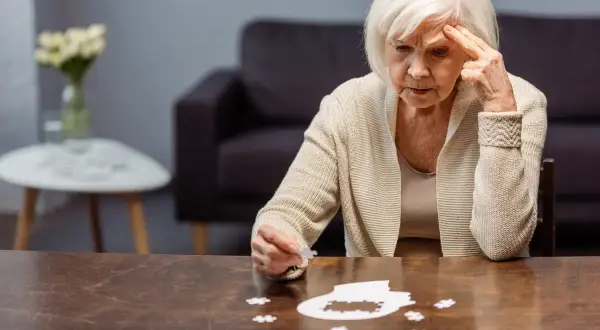 Starsza kobieta zmagająca się z układanką, symbolizującą wczesny alzheimer. Jej zamyślona twarz i gest wskazują na trudności z pamięcią.