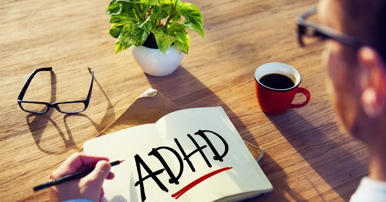Osoba zmagająca się ze spektrum ADHD pisze w zeszycie. Obok leżą okulary, filiżanka kawy i roślina.
