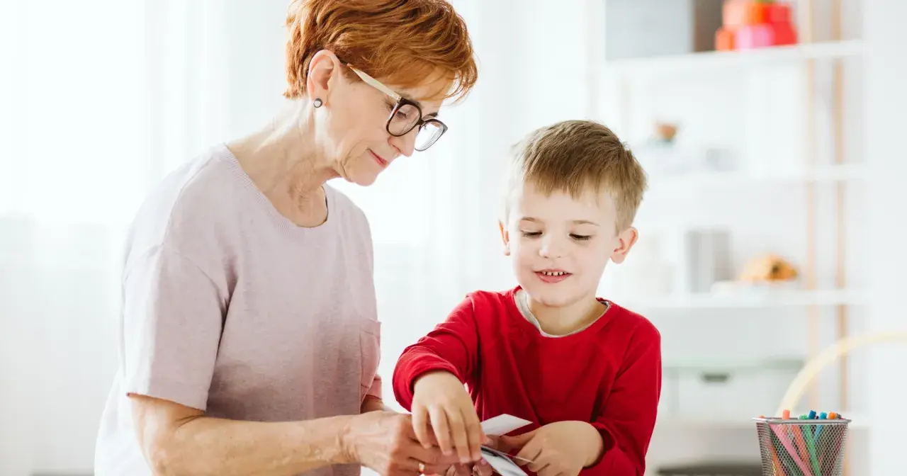 Babcia i wnuczek tworzą coś razem. Dziecko z zaciekawieniem przygląda się swoim ruchom, a jego manieryzmy ruchowe są widoczne w skupieniu.