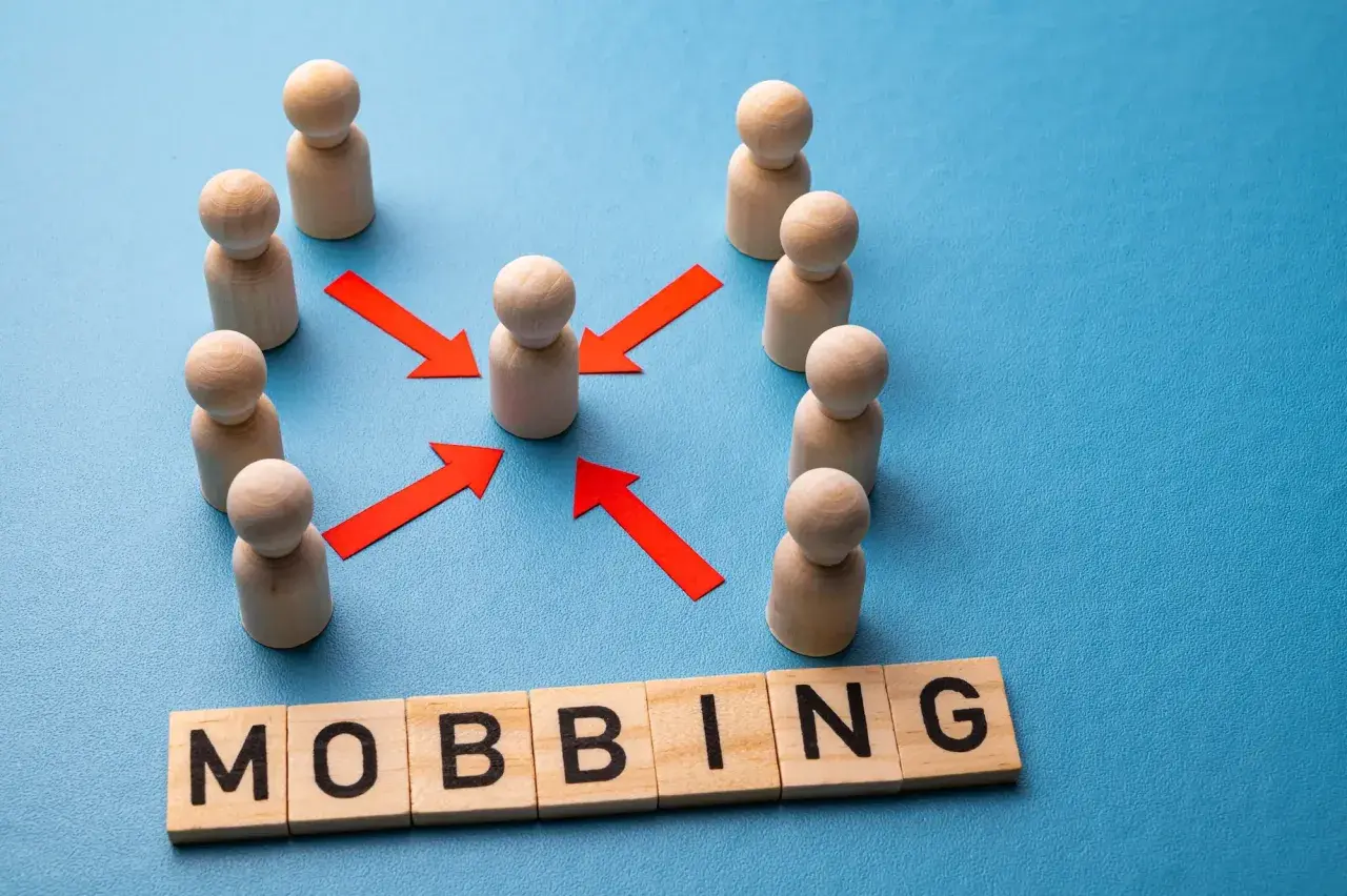 Drewniane ludziki otaczają jednego, a czerwone strzałki wskazują na niego. Pod spodem litery układają się w słowo "MOBBING", symbolizując presję i izolację w miejscu pracy.