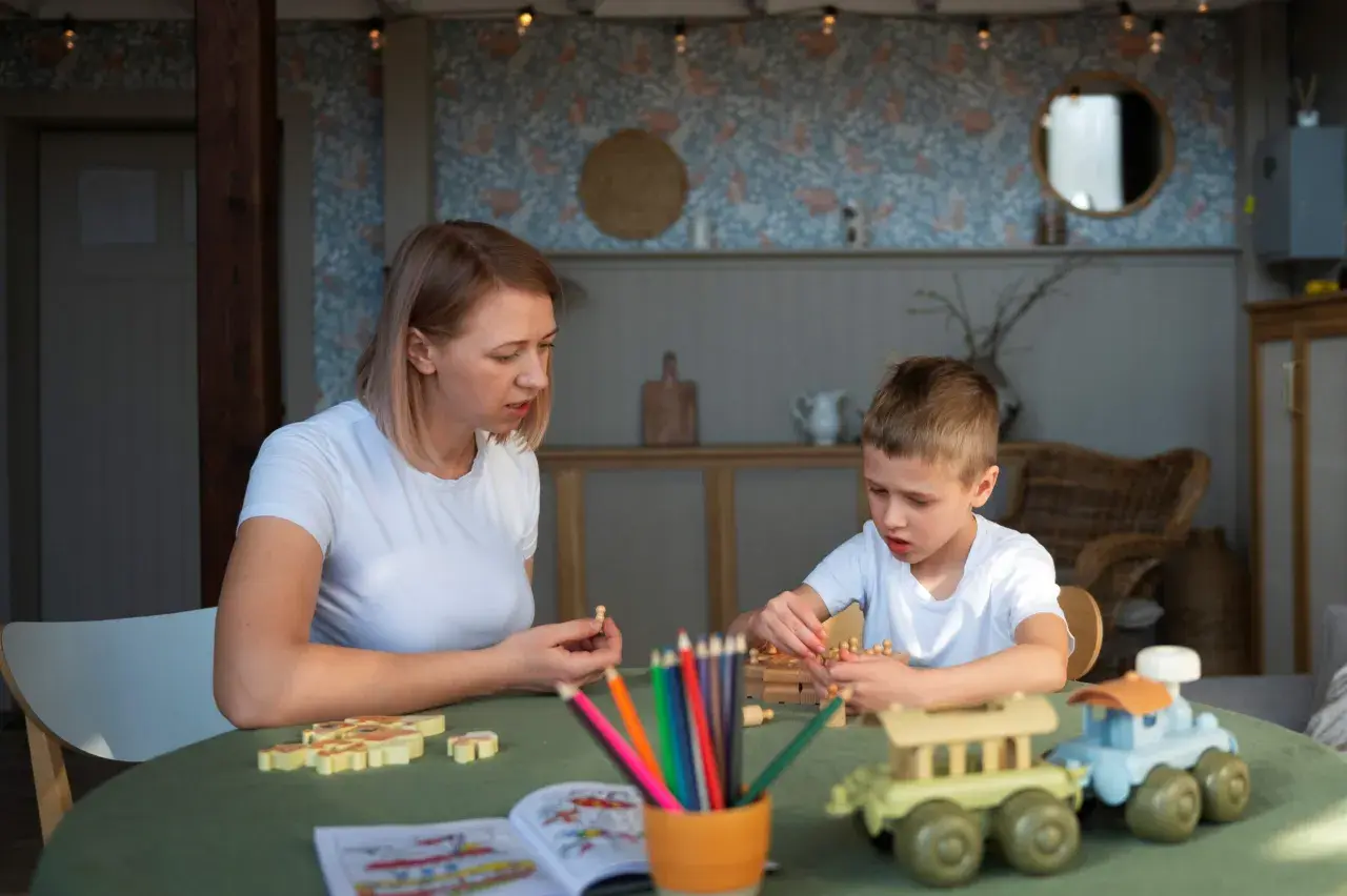 Mama i syn bawią się drewnianymi klockami i kredkami. Tematyka zabawy nawiązuje do przedszkola, rozwijając kreatywność i wyobraźnię.