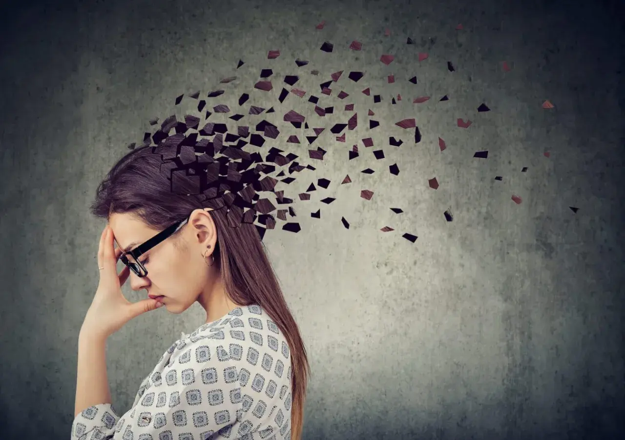 Kobieta z ręką na czole, której głowa rozpada się na fragmenty. Obraz symbolizuje **adhd z przewagą zaburzeń koncentracji uwagi**, trudności w skupieniu myśli.