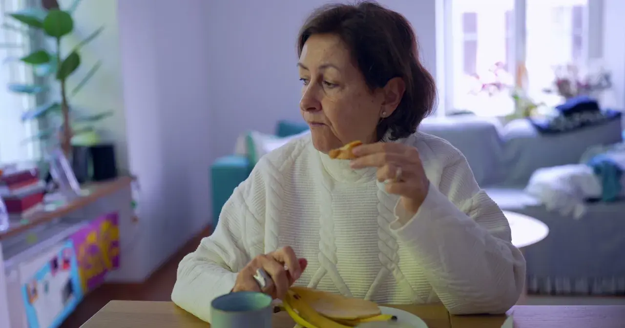Kobieta w białym swetrze je banana, czując uczucie guli w gardle. Na stole kubek i talerz.
