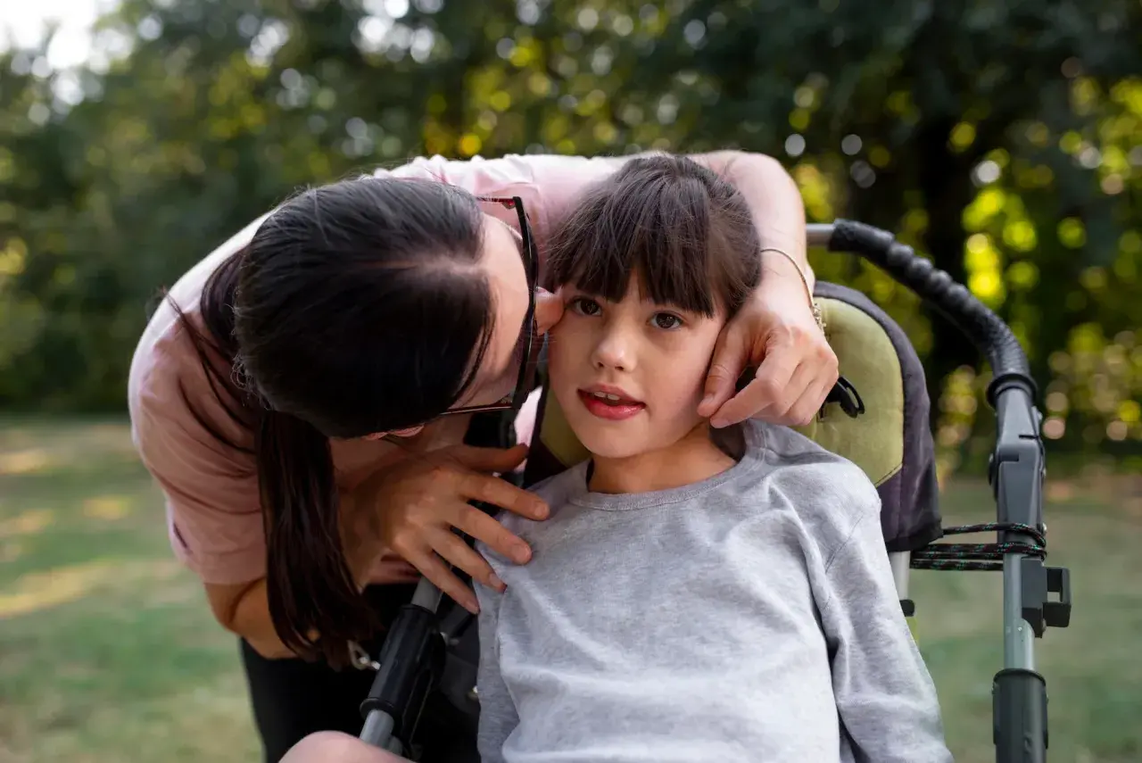 Czuła matka pochyla się nad córką w wózku, troskliwie dotykając jej twarzy. Obraz symbolizuje miłość i wsparcie, a także może nawiązywać do złożonych porażenie mózgowe przyczyny.