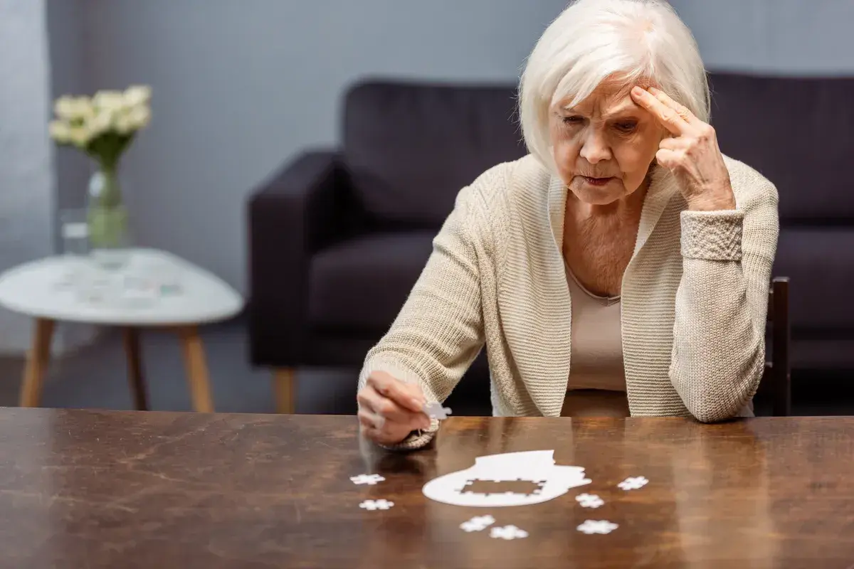 Starsza kobieta zmagająca się z układanką, symbolizującą wczesny alzheimer. Jej zamyślona twarz i gest wskazują na trudności z pamięcią.
