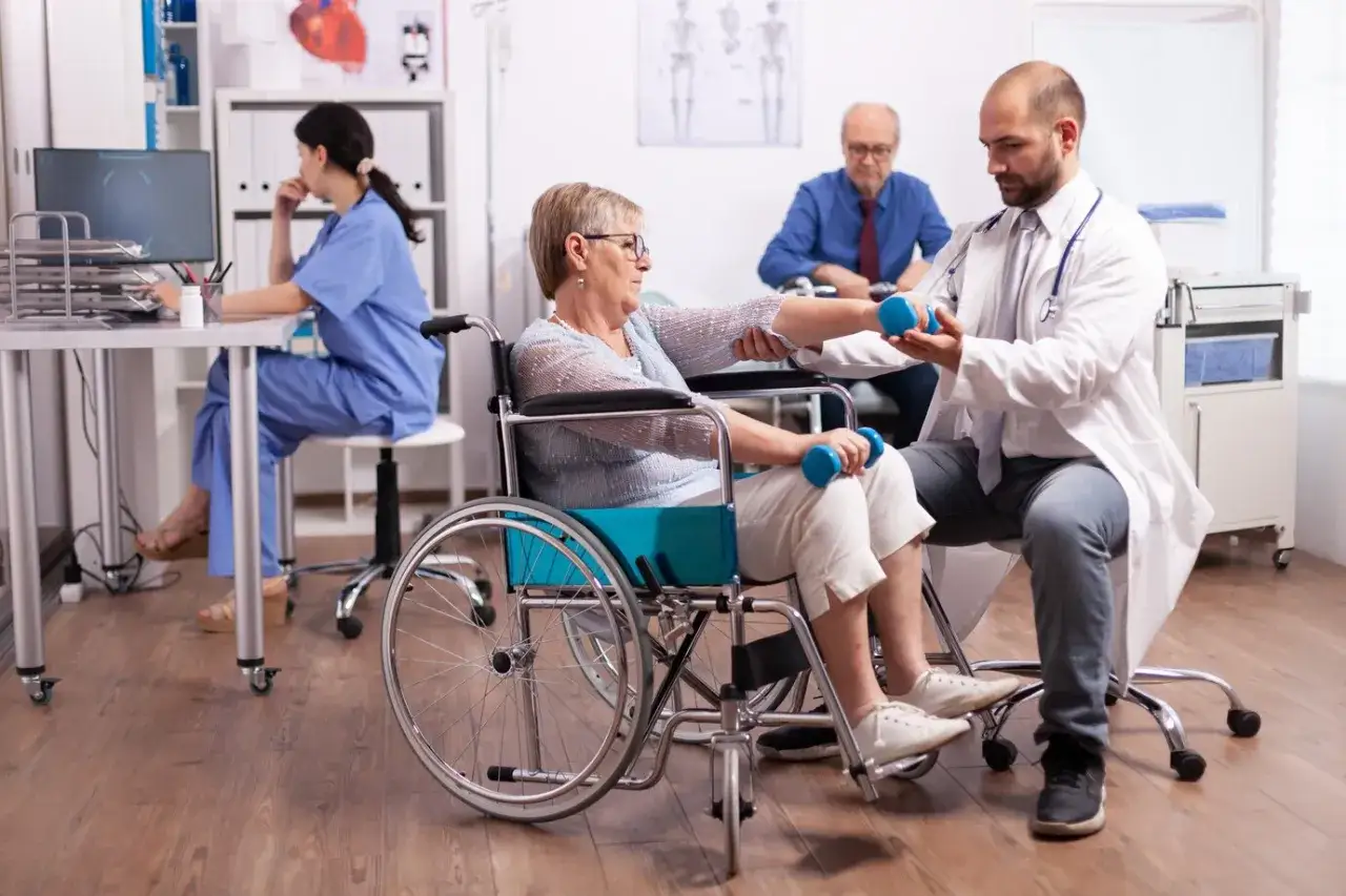 Rehabilitacja po udarze. Kobieta na wózku wykonuje ćwiczenia z hantlami pod okiem fizjoterapeuty.