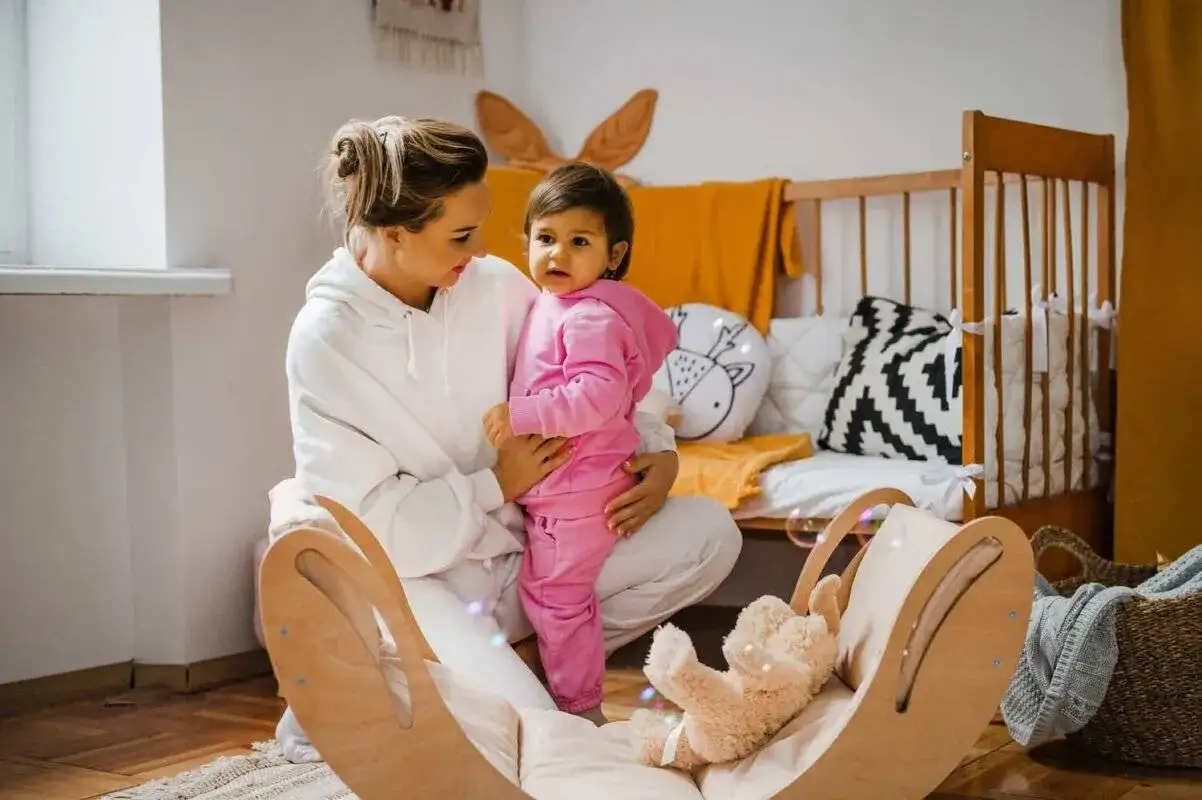 Mama bawi się z dzieckiem, pokazując przykładowe zabawy z dzieckiem autystycznym. W pokoju dziecięcym jest łóżeczko, huśtawka i pluszowy miś.