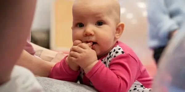 Malutkie dziecko w różowej bluzeczce z serduszkami, z paluszkami w buzi, delikatnie buja się w przód i tył, odkrywając świat.
