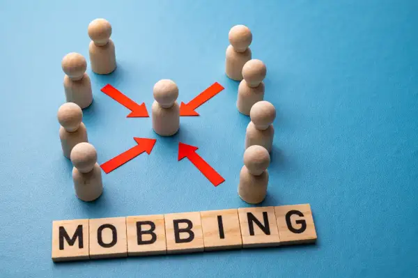 Drewniane ludziki otaczają jednego, a czerwone strzałki wskazują na niego. Pod spodem litery układają się w słowo "MOBBING", symbolizując presję i izolację w miejscu pracy.