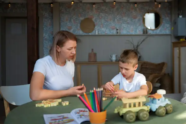 Mama i syn bawią się drewnianymi klockami i kredkami. Tematyka zabawy nawiązuje do przedszkola, rozwijając kreatywność i wyobraźnię.