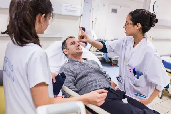 Lekarka bada pacjenta leżącego w łóżku szpitalnym, drugą pielęgniarkę widać z boku.