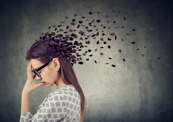 Kobieta z ręką na czole, której głowa rozpada się na fragmenty. Obraz symbolizuje **adhd z przewagą zaburzeń koncentracji uwagi**, trudności w skupieniu myśli.