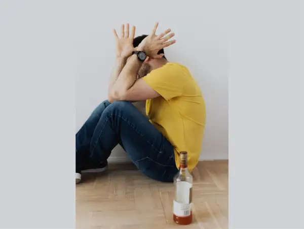 Mężczyzna w żółtej koszulce siedzi na podłodze, zakrywając twarz dłońmi. Obok leży pusta butelka. Symbolizuje trudny proces odstawienia alkoholu.