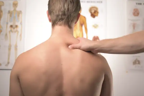 Fizjoterapeuta bada plecy pacjenta, szukając oznak zapalenia rdzenia kręgowego. Na ścianie wiszą anatomiczne plakaty.